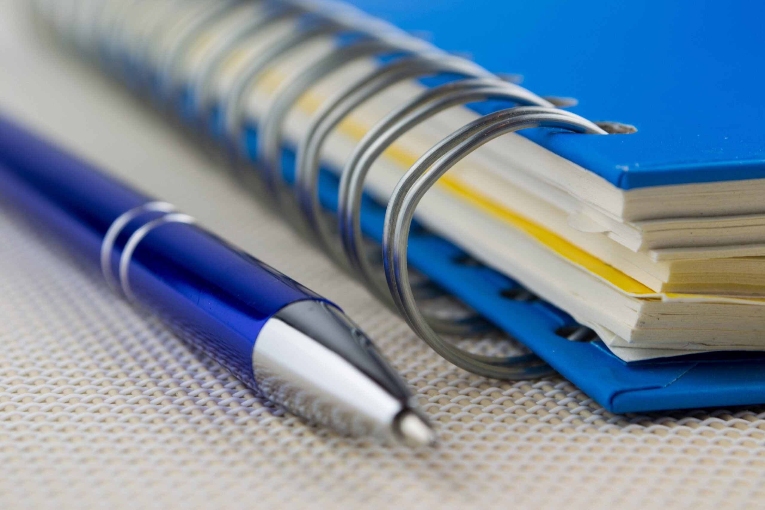 Close-up of a blue pen lying next to a blue spiral-bound notebook to jot down essential nonprofit documents to keep.