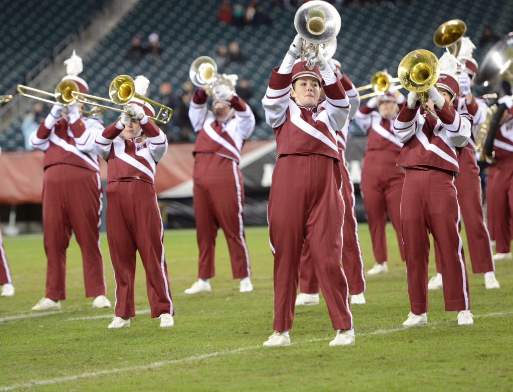 A marching band in red and white uniforms on a football field walking in formation. It's the perfect time to start a booster club to support school activities like music and sports.