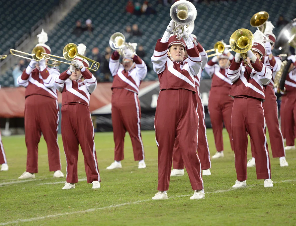 A marching band in red and white uniforms on a football field walking in formation. It's the perfect time to start a booster club to support school activities like music and sports.