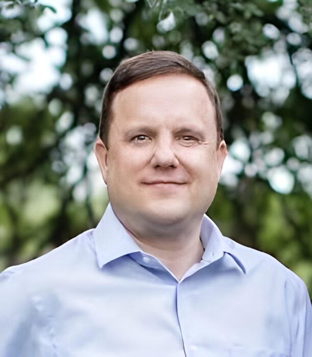 A Texas nonprofit attorney with short brown hair stands outdoors, wearing a light blue collared shirt, with trees in the background.
