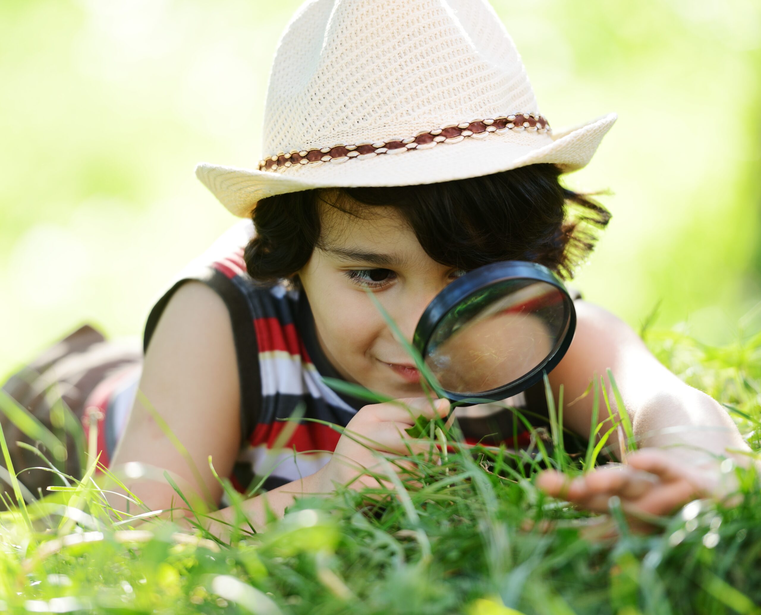 A child uses a magnifying glass to examine the grass while lying on the ground.