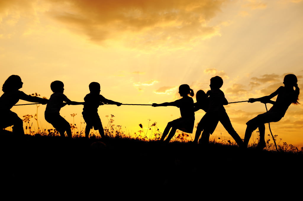 Silhouetted children playing tug-of-war on a hill at sunset with a dramatic sky in the background, symbolizing the dynamic balance of leadership seen between a nonprofit board vs executive director.